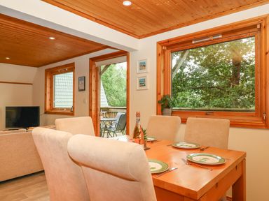 A dining area with a wooden table set for four and beige chairs next to windows and a door leading to a balcony at Lodge 45 - Lazy Bear in Finlakes Holiday Park near Chudleigh