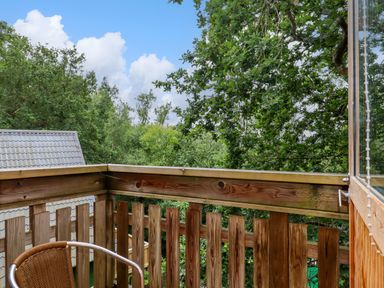 A wooden balcony with a chair overlooking trees and a building with a tiled roof at Lodge 45 - Lazy Bear Finlakes Holiday Park near Chudleigh
