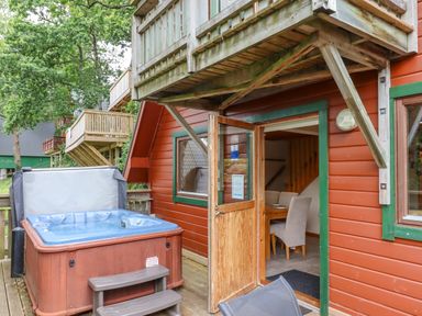 A hot tub on a wooden deck outside a red cabin with an open door showing a dining table and chairs inside at Lodge 45 - Lazy Bear in Finlakes Holiday Park near Chudleigh