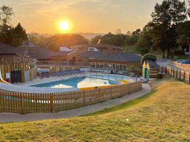 An outdoor swimming pool with a water slide and lounge chairs at sunset surrounded by buildings and trees at Lodge 45 - Lazy Bear in Finlakes Holiday Park near Chudleigh