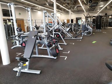 A gym with various weight machines and exercise equipment with black flooring and white ceiling lights