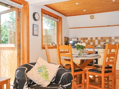 A dining area with a round table set with plates and glasses surrounded by wooden chairs a kitchen with tiled backsplash and an armchair with a plant patterned cushion near a window and door at Lodge 47 - Primrose Lodge in Finlakes Holiday Park near Chudleigh