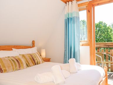 A bedroom with a wooden bed and striped pillows next to a window with blue curtains overlooking a balcony with chairs at Lodge 47 - Primrose Lodge Finlakes Holiday Park near Chudleigh