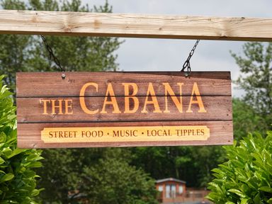A wooden sign hanging from a wooden frame with the words The Cabana street food music local tipples surrounded by green bushes and trees
