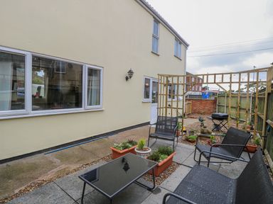 An outdoor patio with black metal chairs a glass table potted plants a wooden trellis and a barbecue grill at Tiffany's Cottage in Gorleston-On-Sea
