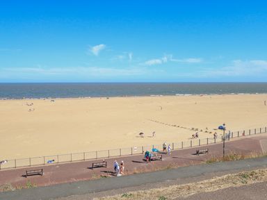 A sandy beach with people walking and sitting on benches near a fenced promenade at Tiffany's Cottage in Gorleston-On-Sea