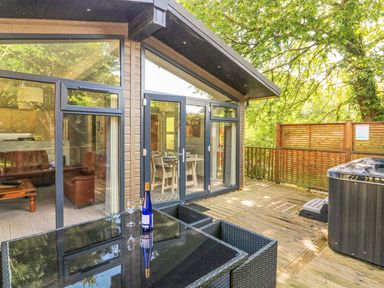An outdoor deck with a glass table set a wine bottle and two glasses a hot tub and patio doors leading to the living area at Woodland View 7 - Oak Cabin Finlakes Holiday Park near Chudleigh