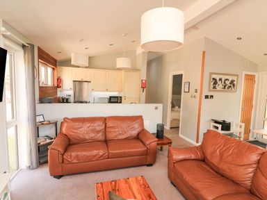 A living room with two brown leather sofas a wooden coffee table and an open kitchen in the background at Woodland View 7 - Oak Cabin Finlakes Holiday Park near Chudleigh