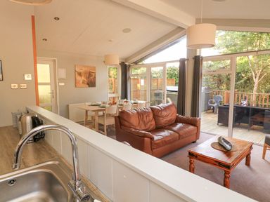 A living room with a brown leather sofa a wooden coffee table and dining table near large windows at Woodland View 7 - Oak Cabin Finlakes Holiday Park near Chudleigh
