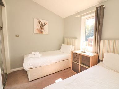 A bedroom with two single beds a small wooden drawer unit with a lamp and a window with curtains at Woodland View 7 - Oak Cabin Finlakes Holiday Park near Chudleigh
