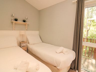 A bedroom with two single beds, a small bedside table with a lamp, a shelf with decorative items, and a window with curtains at Woodland View 7 - Oak Cabin Finlakes Holiday Park near Chudleigh