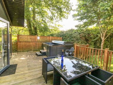 A wooden deck with a hot tub a glass table with a bottle and glasses and outdoor seating at Woodland View 7 - Oak Cabin Finlakes Holiday Park near Chudleigh