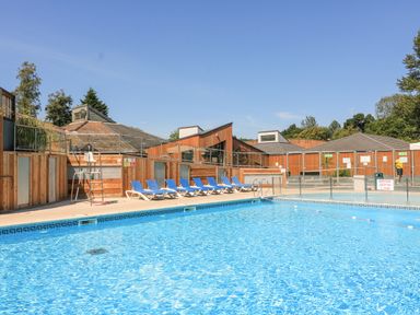An outdoor swimming pool with blue lounge chairs and wooden buildings in the background at Woodland View 7 - Oak Cabin Finlakes Holiday Park near Chudleigh