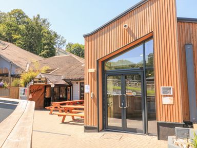An outdoor seating area with wooden picnic tables and buildings including one labeled The Retreat and another with glass doors at Woodland View 7 - Oak Cabin in Finlakes Holiday Park near Chudleigh