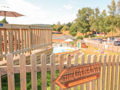 An outdoor area with a wooden fence a swimming pool and a sign pointing to The Cabana with street food music and local tipples at Woodland View 7 - Oak Cabin in Finlakes Holiday Park near Chudleigh