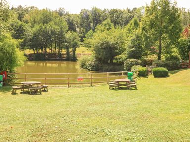 A grassy area with two wooden picnic tables near a fenced pond surrounded by trees at Woodland View 7 - Oak Cabin in Finlakes Holiday Park near Chudleigh