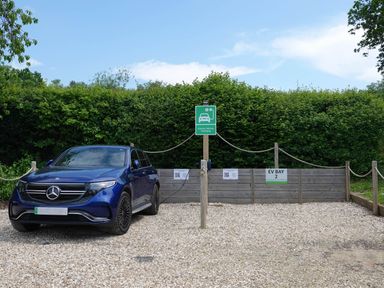 A blue Mercedes electric vehicle parked and charging at EV Bay 2 with a green electric vehicle charging sign and a hedge backdrop at Woodland View 7 - Oak Cabin Finlakes Holiday Park near Chudleigh