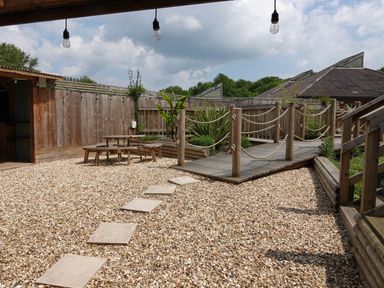 An outdoor area with a gravel ground wooden picnic tables a wooden deck with rope railings and plants at Woodland View 7 - Oak Cabin Finlakes Holiday Park near Chudleigh