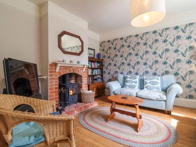 A living room with a blue sofa, wicker chair, wooden coffee table, brick fireplace, and patterned wallpaper at Samphire House in Southwold