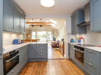A kitchen with blue cabinets wooden floor and white countertops leading to a dining and living area at Samphire House in Southwold