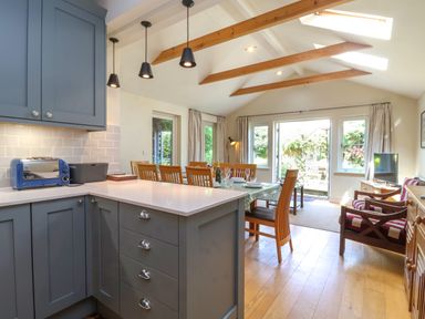A kitchen area with blue cabinets and a toaster next to a dining table with wooden chairs in a room with wooden beams and glass doors at Samphire House in Southwold