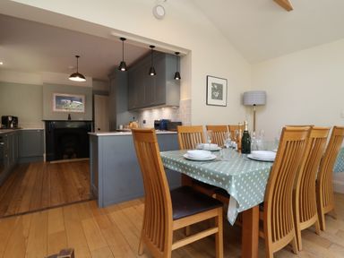 A dining area with wooden chairs and a table set with plates and glasses next to a kitchen with blue cabinets at Samphire House in Southwold
