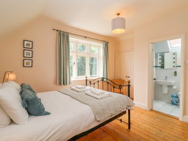 A bedroom with a metal bed frame two folded towels on the bed a window with curtains a wooden chair a lamp and an ensuite bathroom at Samphire House in Southwold