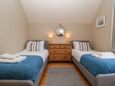 A bedroom with two single beds with blue blankets and striped pillows a wooden dresser with two lamps and a round mirror on the wall a framed picture and a patterned rug on the floor at Samphire House in Southwold