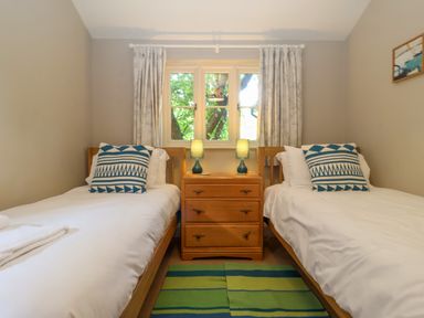 A bedroom with two single beds with pillows a wooden nightstand between the beds with two lamps a window with curtains and a striped rug on the floor at Samphire House in Southwold