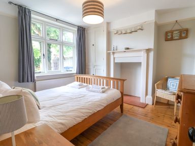 A bedroom with a wooden bed white bedding bedside table with lamp window with curtains wooden floor fireplace wicker chair with cushion Samphire House Southwold