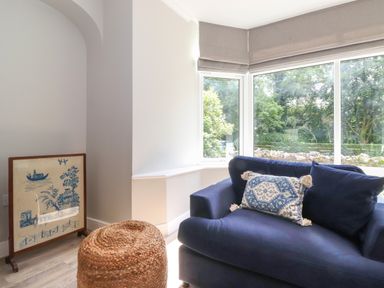 A corner of a room with a blue sofa and a patterned cushion near large windows with beige blinds and a woven round ottoman next to a framed artwork on the floor at Eller Cottage in Windermere