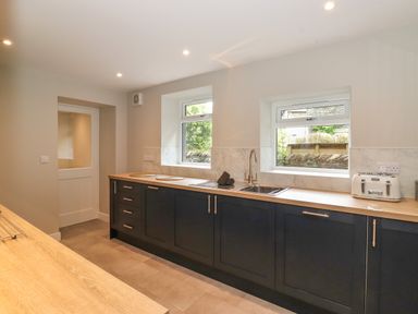 A kitchen with dark lower cabinets a wooden countertop a double sink two windows and a white door at Eller Cottage in Windermere