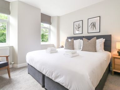 A bedroom with a large bed with pillows and folded towels beside two windows and a wooden nightstand with a lamp at Eller Cottage in Windermere