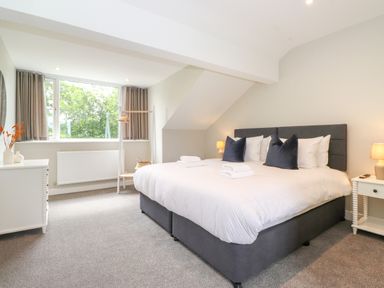 A bedroom with a large bed with white linens and dark pillows two bedside tables with lamps a window with curtains and a clothing rack at Eller Cottage in Windermere