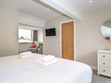 A bedroom with a bed, towels, a wooden door, a wall-mounted TV, a white dresser, and a seating area near a window at Eller Cottage in Windermere