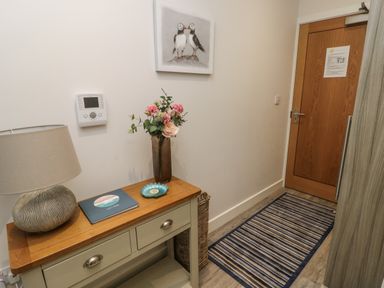 A hallway with a striped rug a wooden door a table with drawers a lamp a vase with flowers and a guest book Sandy Bay Retreat Benllech