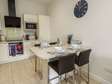 A kitchen with a dining table set for four with chairs a clock on the wall and kitchen appliances at Sandy Bay Retreat in Benllech