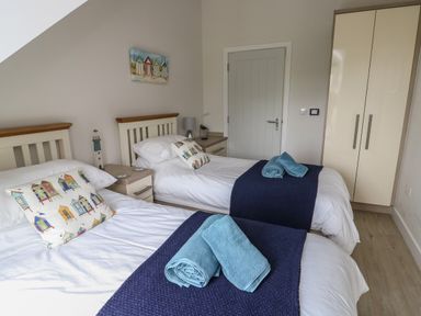 A bedroom with two single beds each with white bedding pillows with beach hut patterns navy blue runners and blue towels a nightstand between beds a white door and a beige wardrobe on light wood flooring at Sandy Bay Retreat in Benllech