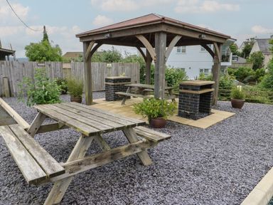 An outdoor area with wooden picnic tables a wooden gazebo and potted plants on a gravel surface at Sandy Bay Retreat in Benllech