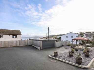 An outdoor area with a paved surface a wooden fence potted plants a wooden gazebo picnic tables and houses in the background at Sandy Bay Retreat in Benllech