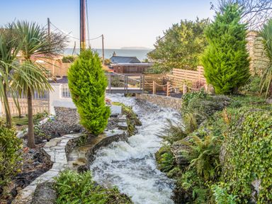 A garden with a flowing stream stone pathway wooden bridge and trees with the sea in the background at Sandy Bay Retreat in Benllech