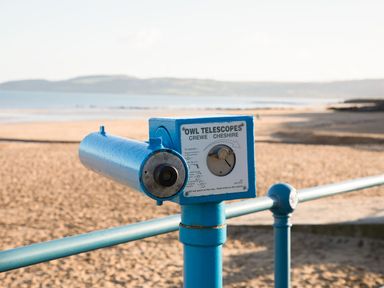 A blue coin-operated viewing telescope on a beach with sea and hills in the background at Sandy Bay Retreat in Benllech