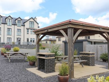 An outdoor seating area with wooden picnic tables under a wooden gazebo and potted plants near residential buildings at Sandy Bay Retreat in Benllech