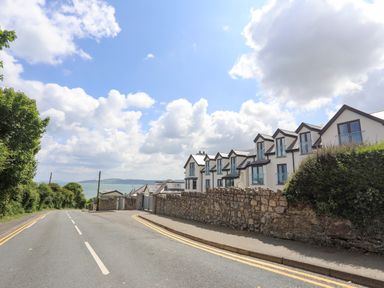 A road with a stone wall and modern houses on the right and trees on the left with a view of the sea at Sandy Bay Retreat in Benllech