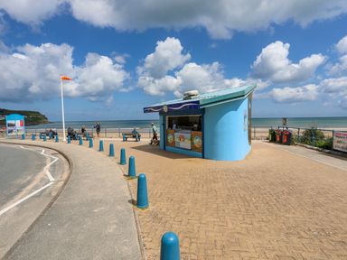 A blue kiosk on a paved area near the beach with people sitting on benches and a sea view at Sandy Bay Retreat in Benllech