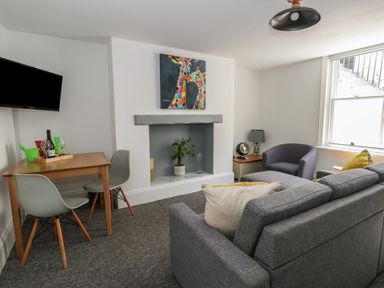 A living room with a gray sofa, two chairs, a small wooden dining table with two chairs, a wall-mounted television, a fireplace with a plant inside, and a colorful giraffe painting above at Flat 1 - Sandbanks in Bournemouth