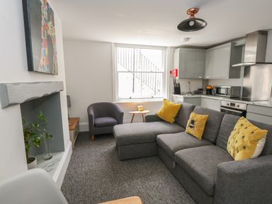 A living room with a grey sofa, yellow cushions, a grey armchair, a small wooden table, a fireplace with a plant, and a kitchen area at Flat 1 - Sandbanks in Bournemouth