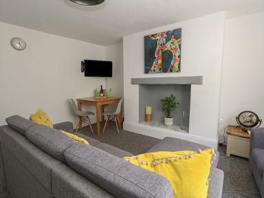 A living room with a gray sofa with yellow cushions a wooden table with two gray chairs a television mounted on the wall a decorative fireplace with a plant a candle and a vase and a painting of colorful giraffes above the fireplace at Flat 1 - Sandbanks in Bournemouth