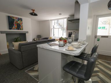 A kitchen with a breakfast bar and black chairs opening into a living room with a grey sofa and colorful giraffe painting on the wall at Flat 1 - Sandbanks in Bournemouth