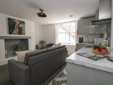 A living room with a grey sofa, armchair, colorful giraffe painting, and a kitchen counter with fruit bowl at Flat 1 - Sandbanks in Bournemouth